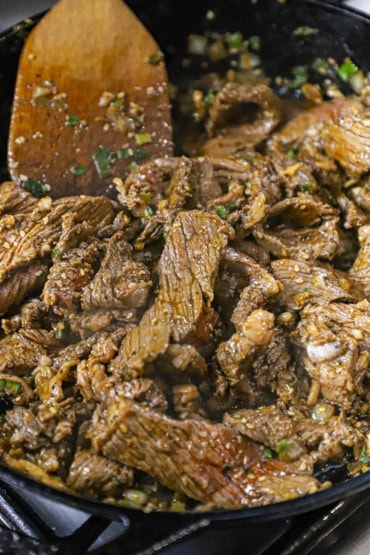 A close-up view of a person using a wooden spatula to stir-fry strips of steak in a large black cast-iron skillet with a Korean sauce and marinade.