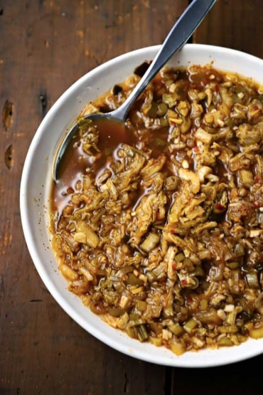 An overhead view of a white bowl that is holding homemade kimchi with a spoon inserted into the side.