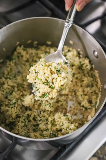 An overhead view of a person holding a serving spoon filled with a serving of cooked brown rice with cilantro and lime in a medium-sized saucepan on a gas stove.