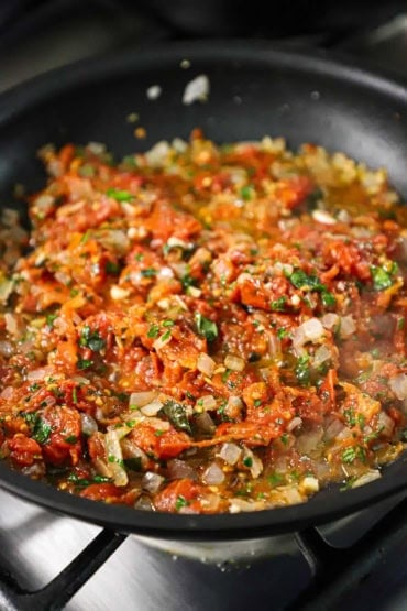 A straight-on view of a large black non-stick skillet filled with a simmering Mediterranean tomato, onion, and black olive sauce.
