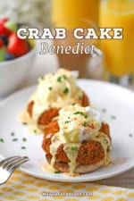 A straight-on view of two crab cake Benedict servings on a single white dinner plate with a tall glass of orange juice and a bowl of fruit in the background