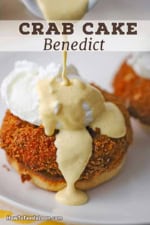 A close-up view of hollandaise sauce being poured from a small white pitcher onto a poached egg that is resting on top of a crispy crab cake and the bottom of an English muffin.