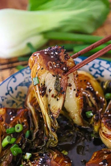 A close-up view of a sautéed baby bok choy being raised with chopsticks from a bowl of the same with sesame seeds sprinkled over the top.