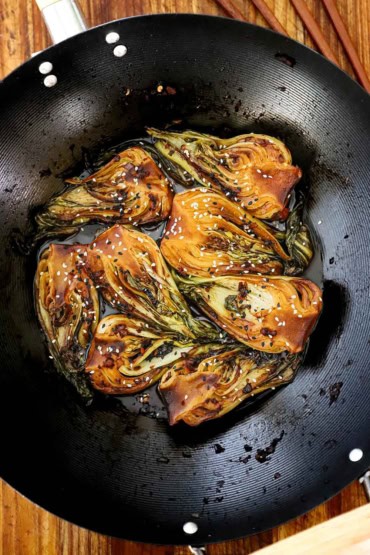 An overhead view of four baby bok choy that have been cut in half lengthwise and have been sautéed in a dark sauce in a black wok.