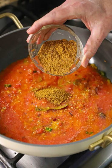 A person transferring homemade chana masala powder from a small glass bowl into a large deep skillet filled with simmering puréed tomatoes, onions, and spices.