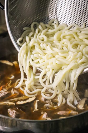 A close-up view of cooked udon noodles being transferred from a metal colander into a large skillet filled with a simmering beef broth with mushrooms and slivers of carrots.