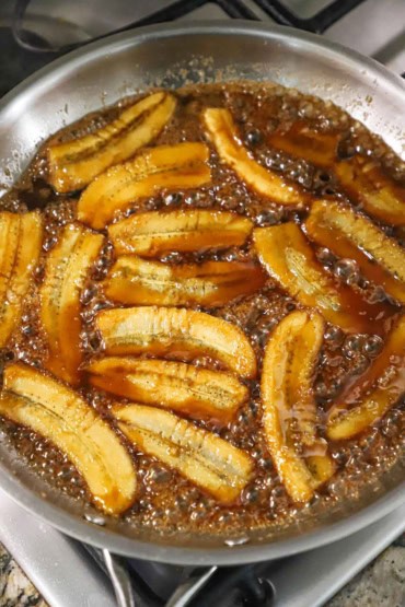 An overhead view of a large stainless steel skillet filled with a layer of quartered bananas simmering in a thick caramel sauce on the stove.