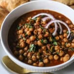 A close-up view of a white bowl holding a serving of chana masala topped with chopped cilantro and two slivers of red onion and a gold spoon next to the bowl.
