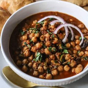 A close-up view of a white bowl holding a serving of chana masala topped with chopped cilantro and two slivers of red onion and a gold spoon next to the bowl.