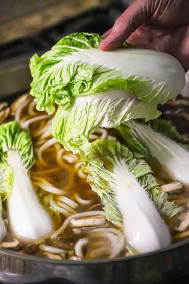 A person placing Chinese cabbage leaves into a large skillet that is filled with a simmering beef broth, udon noodles, and sliced mushrooms.
