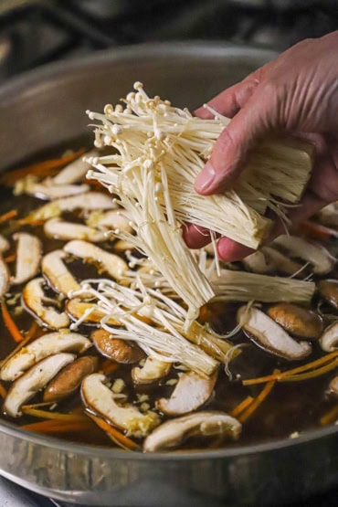 A person placing a handful of uncooked enoki mushrooms into a large skilet filled with simmering beef broth with sliced shiitake mushrooms, ginger, and carrot slivers.