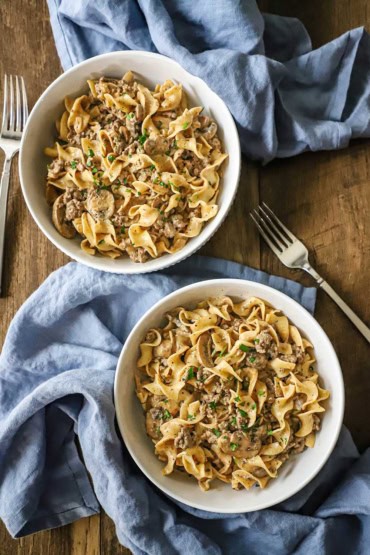 An overhead view of two white bowls each filled with a serving of weeknight beef stroganoff with blue napkins nearby as well as two silver forks.