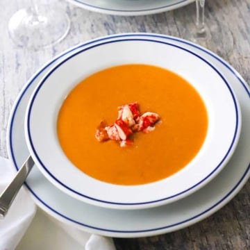 A close-up view of a bowl of lobster bisque in a shallow white bowl with a blue thin-line rim and there is fresh lobster chunks resting on top.