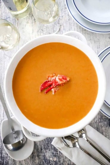 An overhead view of a large white soup terrine filled with lobster bisque with a meat from a lobster claw resting in the middle of the soup.