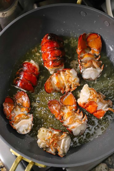 An overhead view of four lobster tails that have been cut in half and are being cooked in bubbling butter in a large deep skillet.
