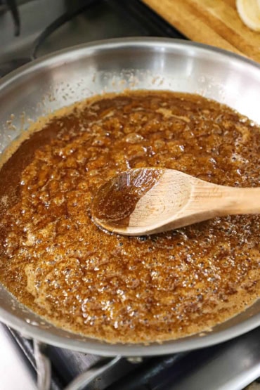 A straight-on view of a large silver skillet filled with a simmering brown sugar and butter sauce that has a large wooden spoon resting the middle.