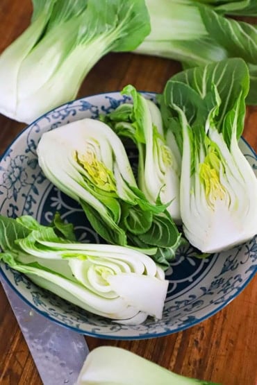A straight-on view of two fresh baby bok choy cabbages that have been split in half, lengthwise and are resting in a colorful bowl in a cutting board next to a knife.