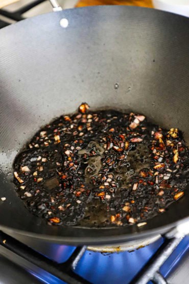 A close-up view of a wok that is over a gas flame on stove with a simmering dark sesame oil with garlic and ginger sauce in the bottom in it.