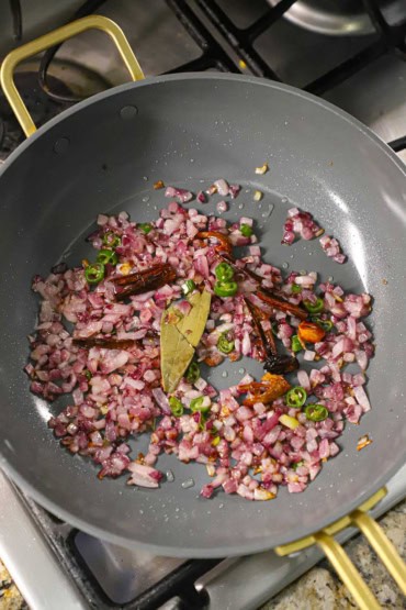 An overhead view of a large, deep skillet filled with chopped red onions, a bay leaf, several cinnamon sticks, mace, and chopped green pepper being sautéed.