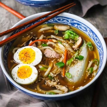 A straight-on view of a large shallow colorful bowl filled with udon noodle soup with pork including two halves of hard boiled eggs and Chinese cabbage in the soup.
