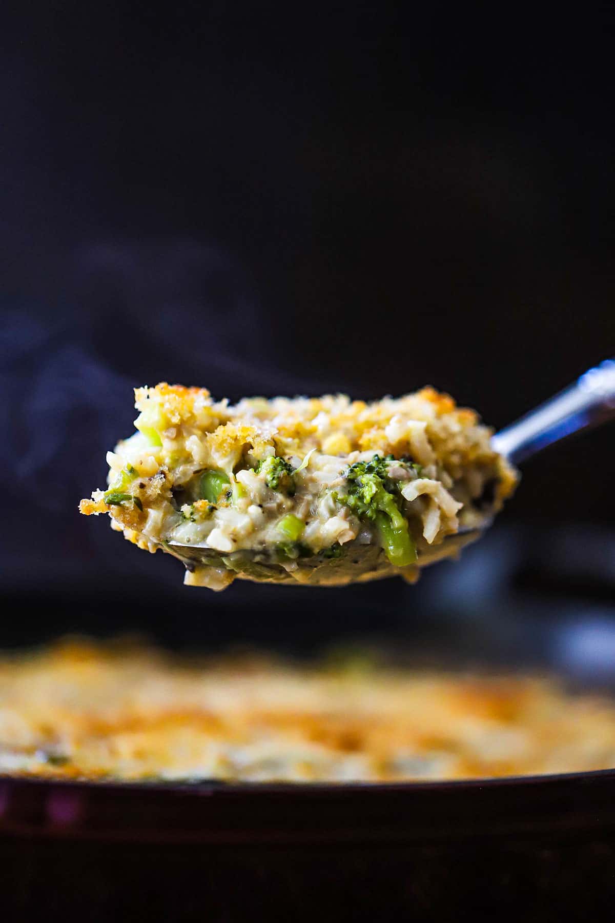 A large serving spoon lifting up a helping of broccoli casserole with cheddar and rice over a dish of the same.