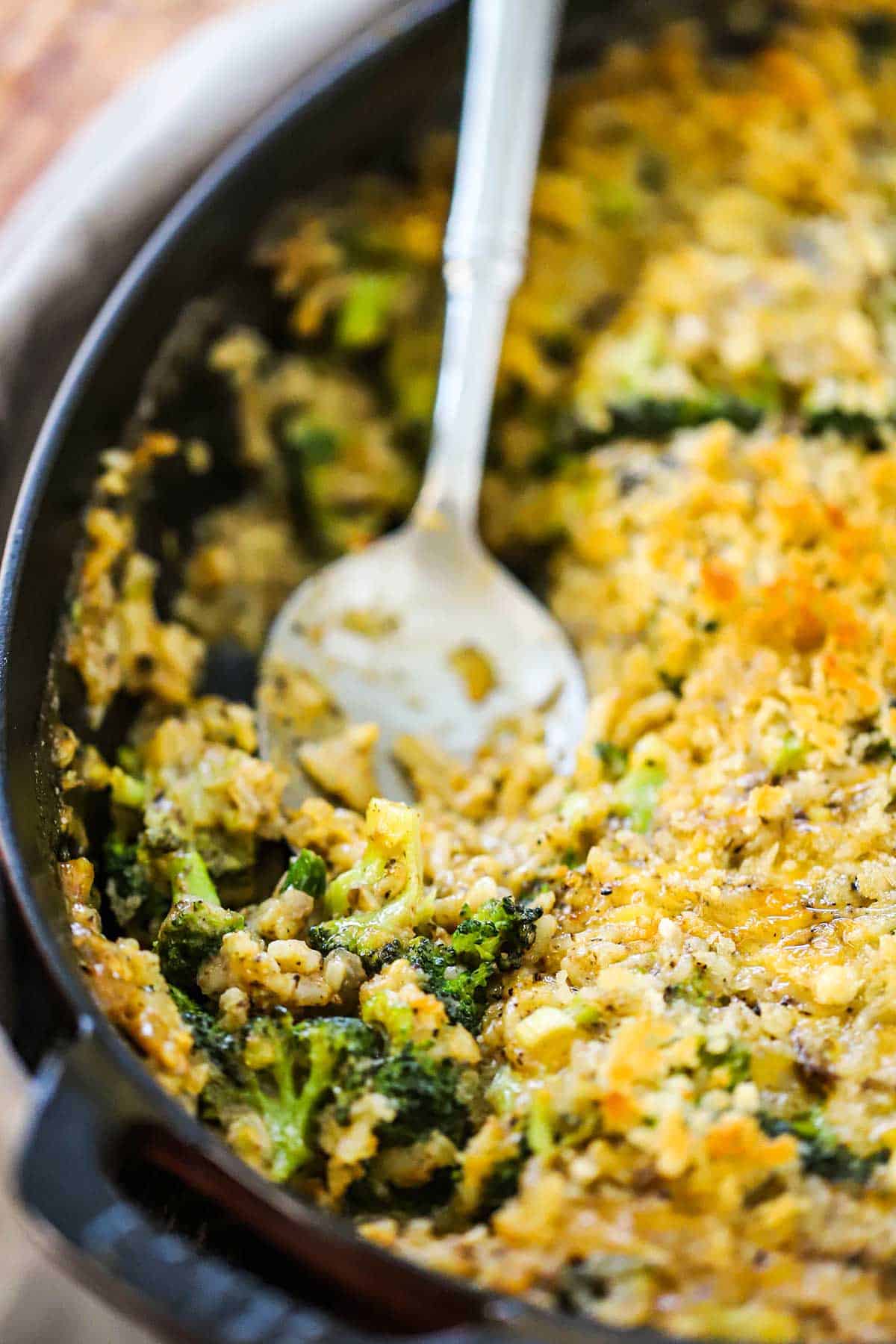 A large silver serving spoon inserted into a dish holding broccoli casserole with cheddar and rice.