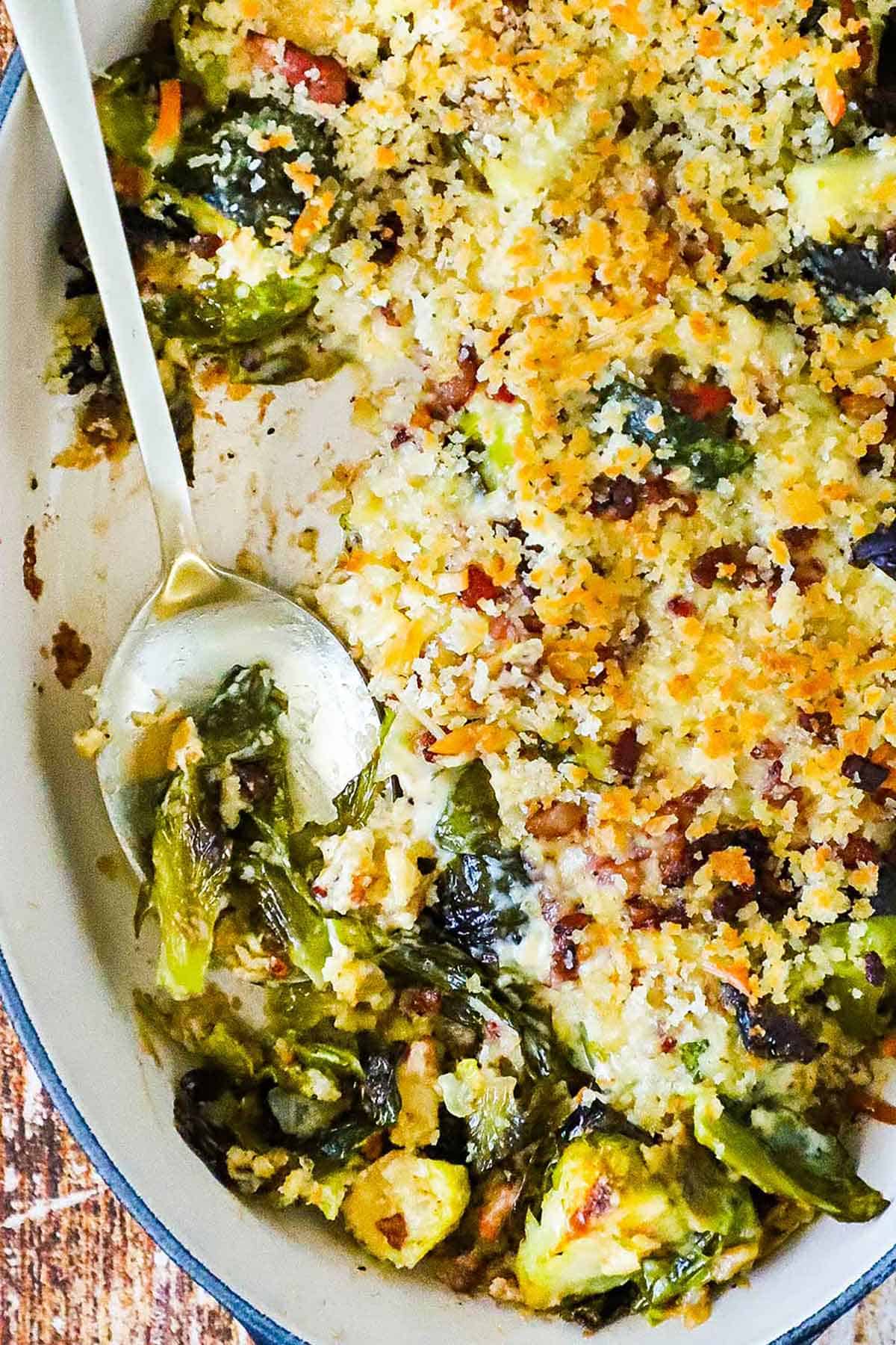 An oval baking dish filled with Brussels sprouts gratin topped with toasted breadcrumbs with a spoon in the side.