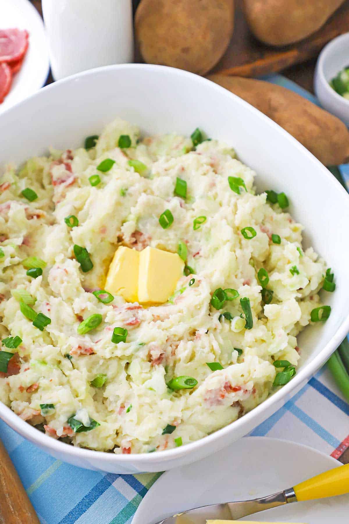 A straight-on view of a large white serving bowl filled with colcannon (Irish mashed potatoes) topped with two melty tabs of butter and garnished with chopped scallions.