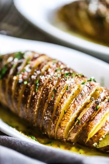 A crispy hasselback potato that is topped with grated parmesan cheese and finely chopped parsley resting in an individual oval baking dish.