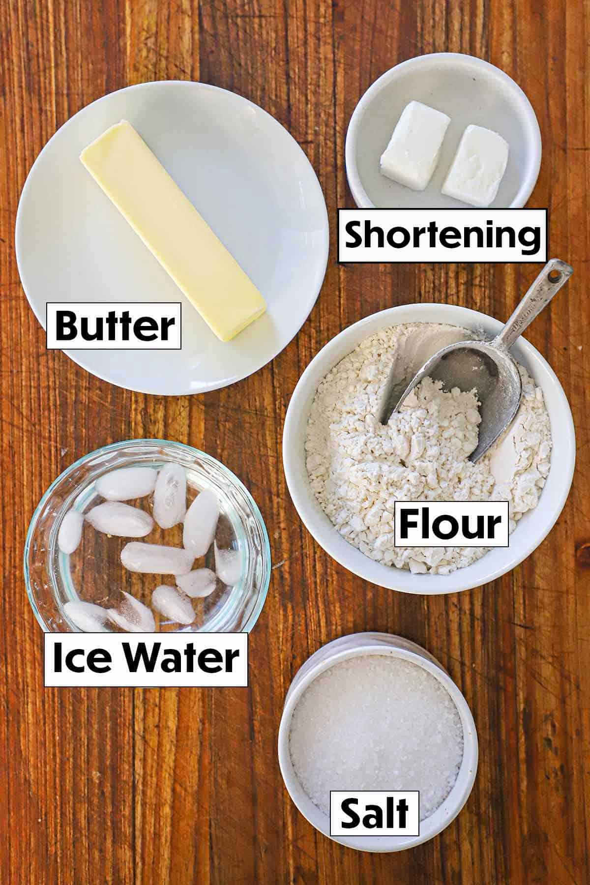 An organized display of ingredients for easy pie crust on a rustic wooden background, featuring all-purpose flour, cold butter, salt, shortening, and a glass of ice water, highlighting their textures and colors.