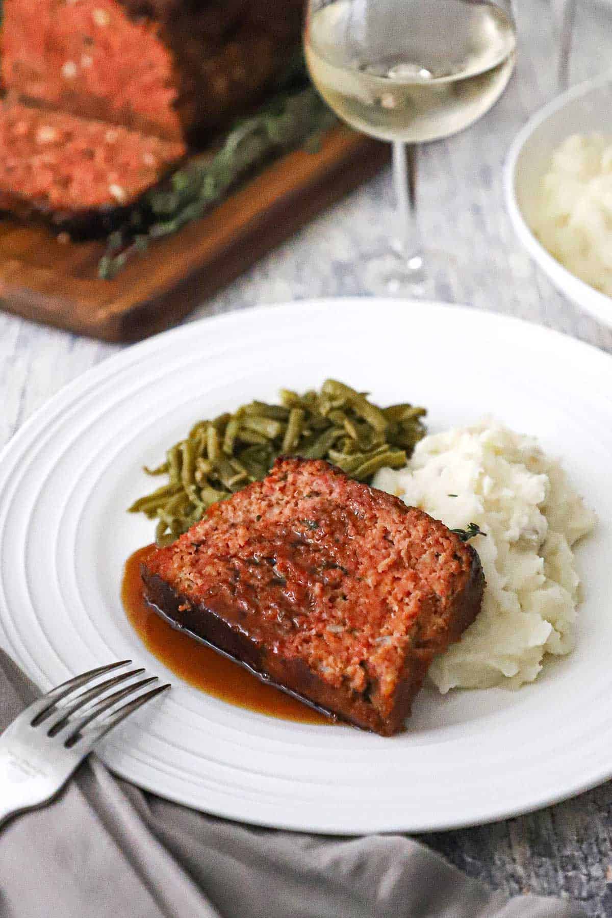 A white dinner plate filled with a serving of slized glazed ham loaf on a pile of mashed potatoes next to Southern-style green beans.
