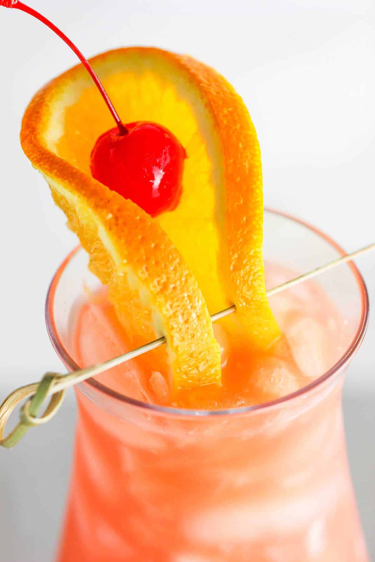 A close-up view of a hurricane cocktail with a folded orange slice that is pierced with a toothpick and a cherry resting on the top.