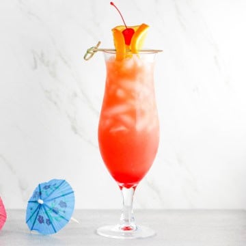 A close-up view of a hurricane cocktail with a blue drink umbrella sitting next to the classic glass.