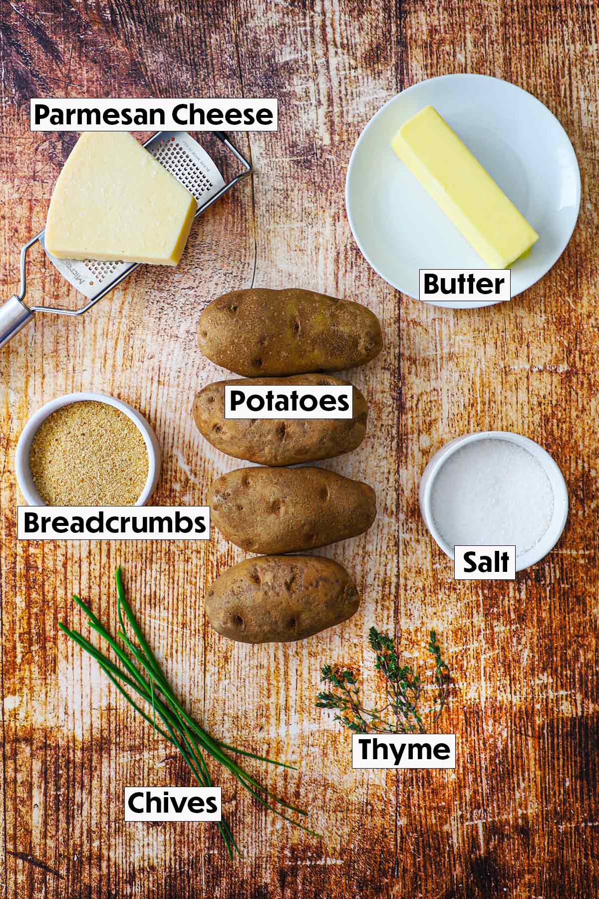 An arrangement of ingredients for Hasselback potatoes on a wooden background including a block of romano cheese, a stick of butter, breadcrumbs, russet potatoes, and fresh herbs.