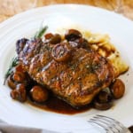 A serving of pork chop agrodolce with mushrooms on a white dinner plate with a glass of white wine next to it.