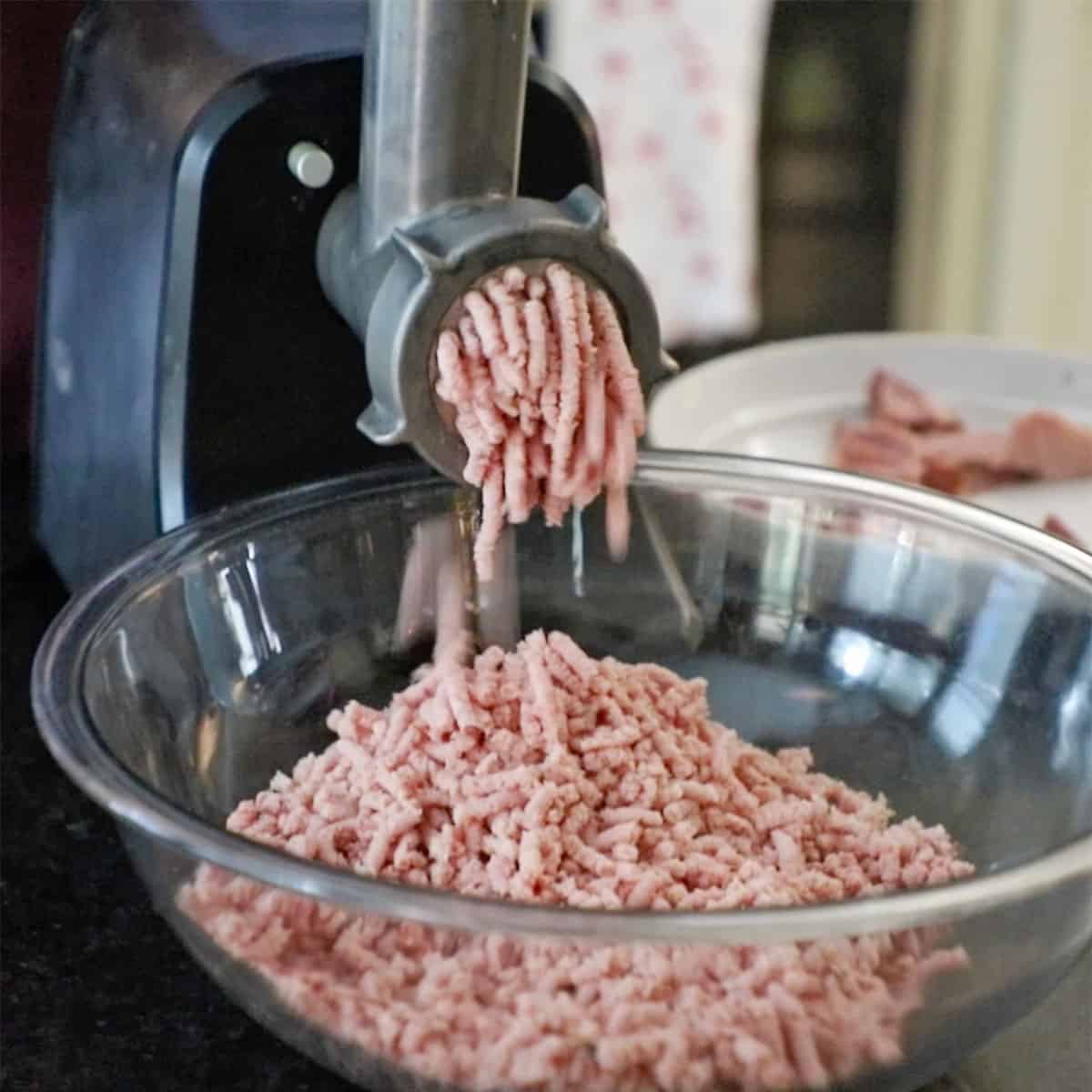 Ground ham exuding from a meat grinder into a glass bowl.