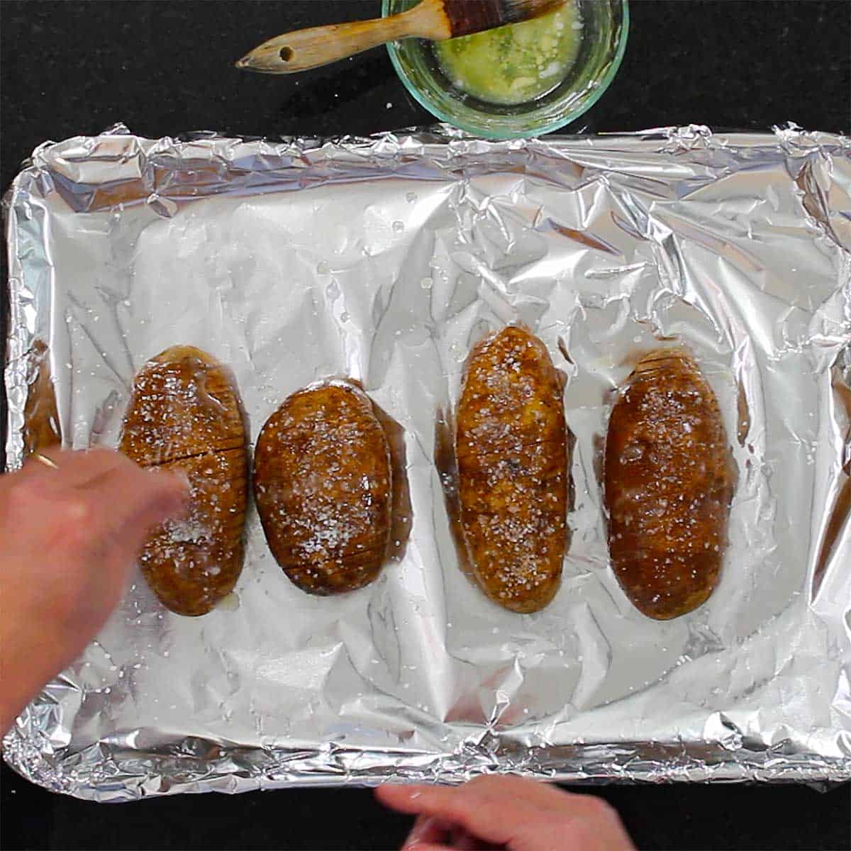 A person sprinkling the tops of sliced potatoes that are resting on a foil-lined baking sheet.