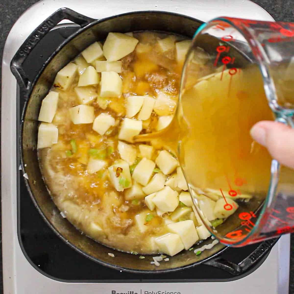 A person pouring chicken broth from a large glass measuring cup into a black pot filled with cubed potatoes.