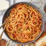 A large, deep skillet filled with Bucatini all’Amatriciana with a block of cheese nearby.