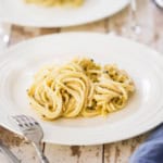 A white dinner plate filled with a serving of tonnarelli cacio e pepe with a glass of white wine nearby.