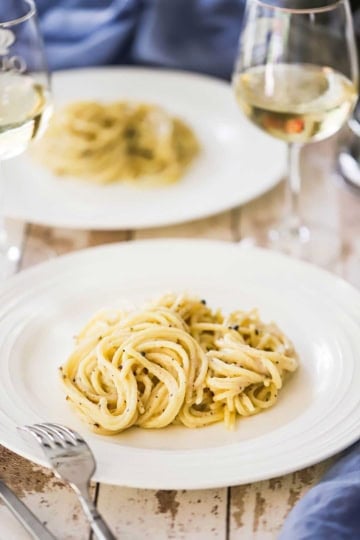 A white dinner plate filled with a serving of tonnarelli cacio e pepe with a glass of white wine nearby.
