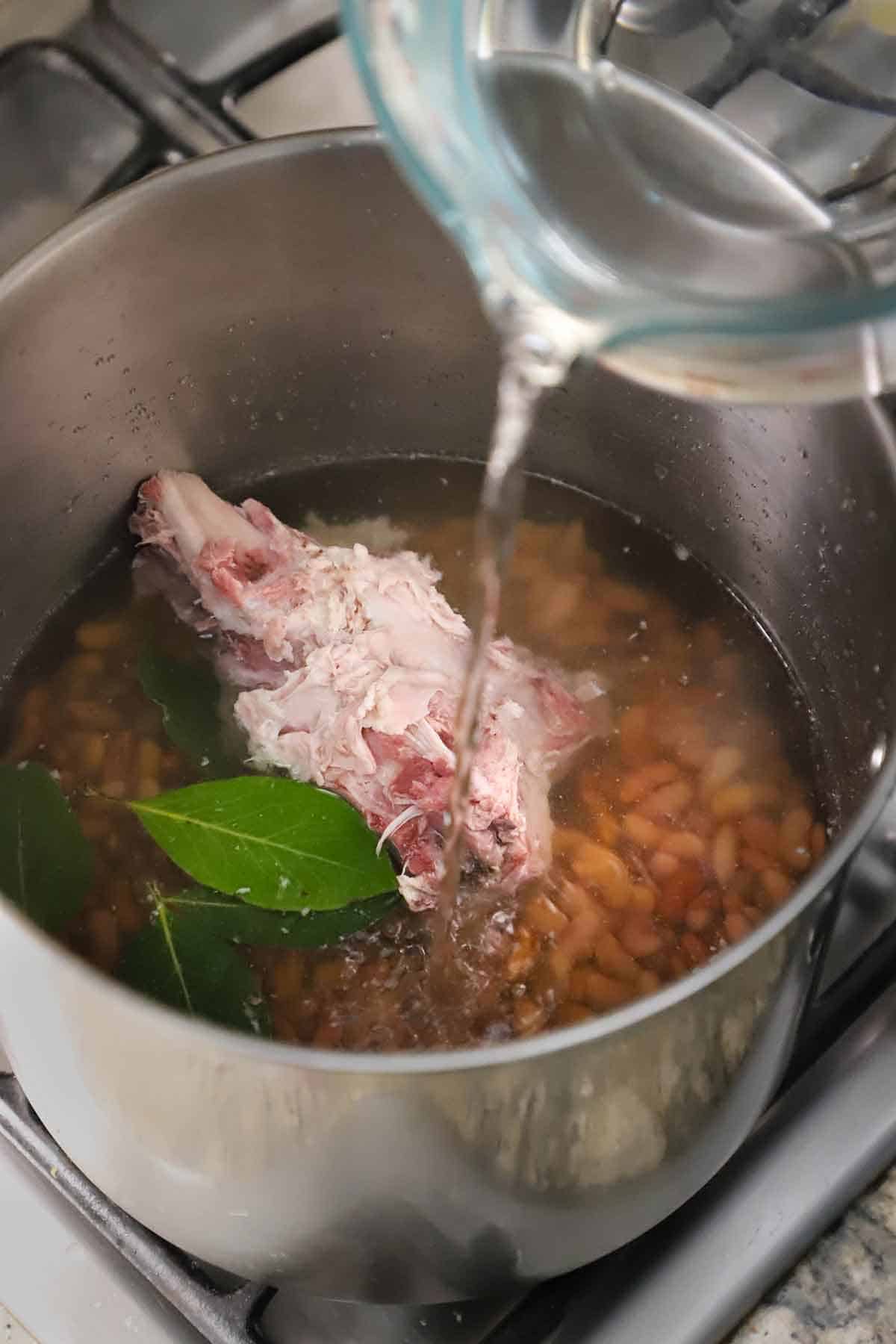 An overhead view of water being poured from a large glass measuring cup into a large stock pot filled with kidney beans, a ham bone, bay leaves, and more water.