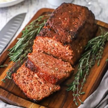 A glazed ham loaf sitting on a small cutting board and has a few slices cut sitting next to sprigs of thyme and rosemary.