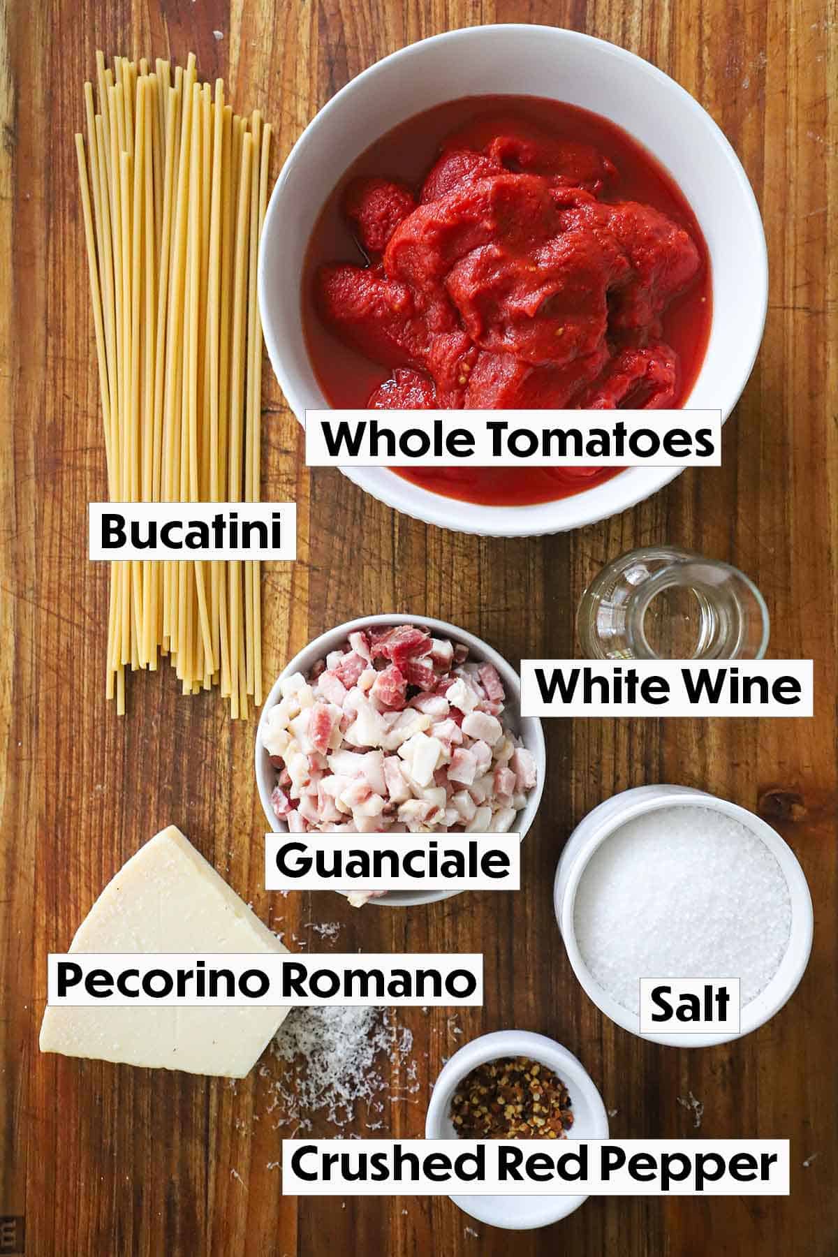 A collection of ingredients for Bucatini all’Amatriciana
including tomatoes, bucatini, guanciale, wine, salt, red pepper flakes, and cheese on a brown cutting board.