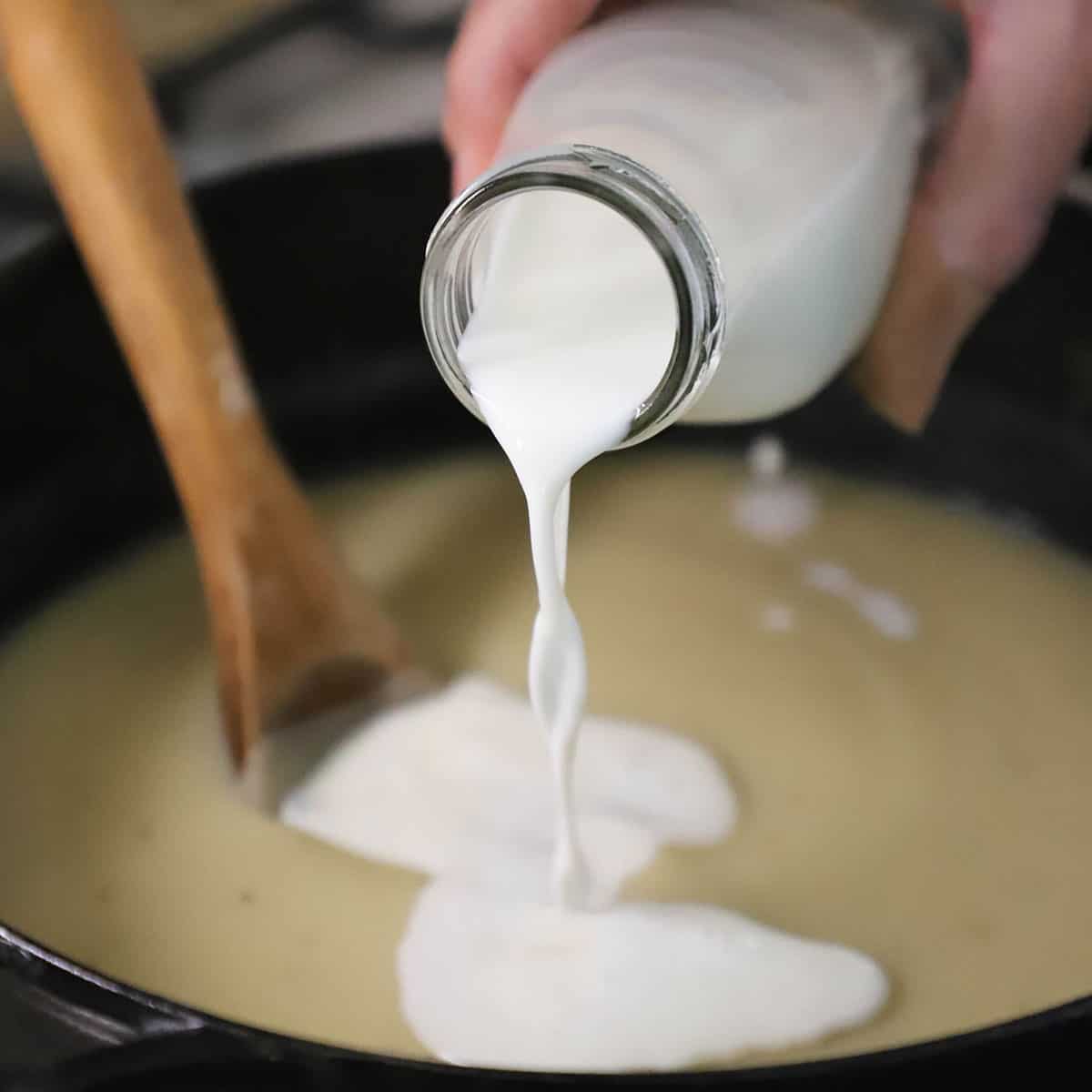 A person pouring whole milk from a small glass milk jug into a pot of puréed Russet potatoes.