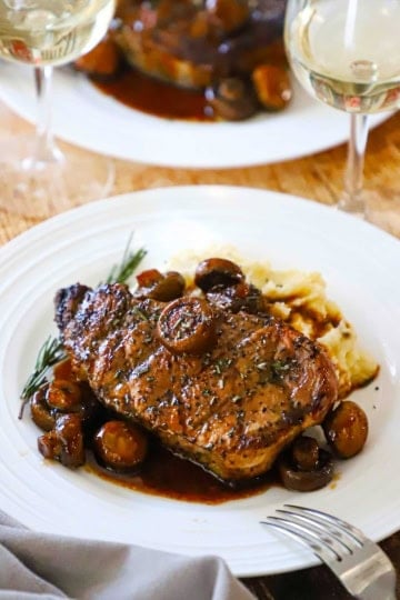 A serving of pork chop agrodolce with mushrooms on a white dinner plate with a glass of white wine next to it.