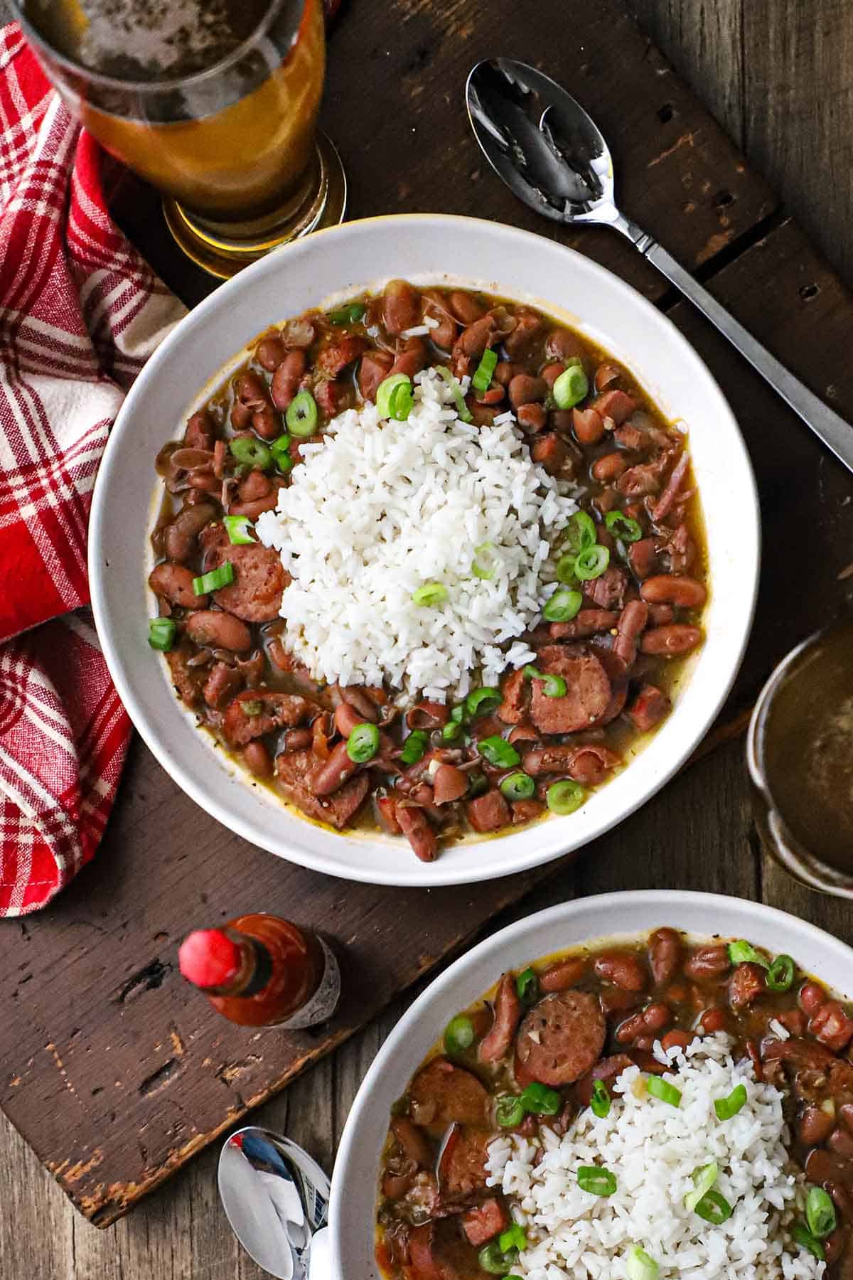 An overhead view of two white bowls filled with servings of red beans and rice with a small bottle of hot sauce and a beer in a tall glass nearby.