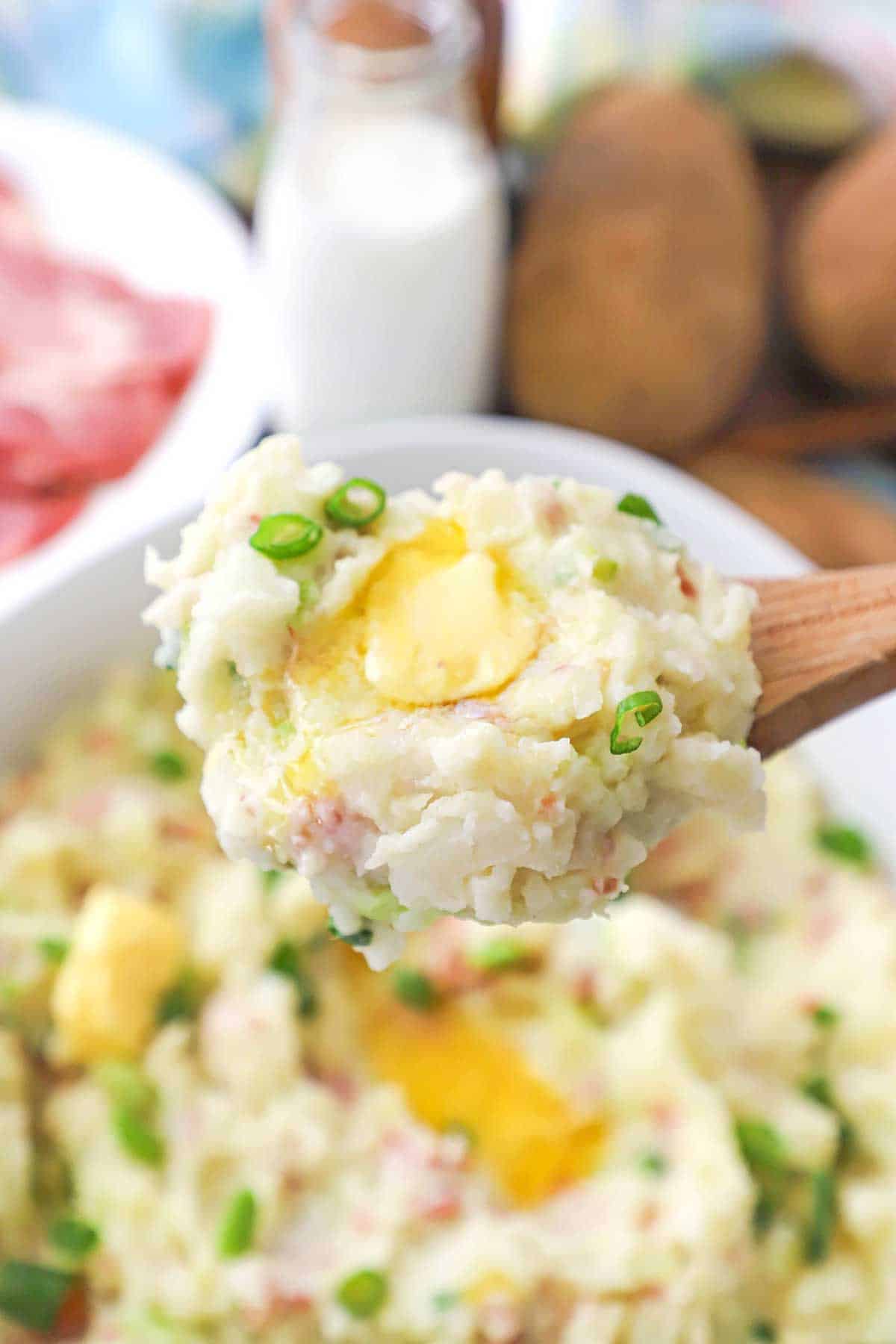 A close-up view of a wooden spoon being used to hold up a large serving of colcannon (Irish mashed potatoes) with a pad of melting butter on top.