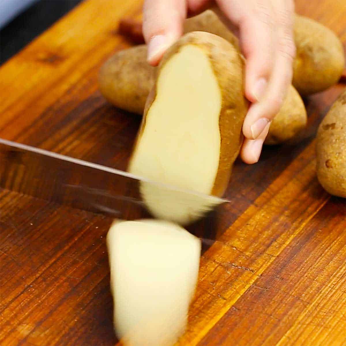 A person using a large knife to slice a thin strip from the side of a russet potato.