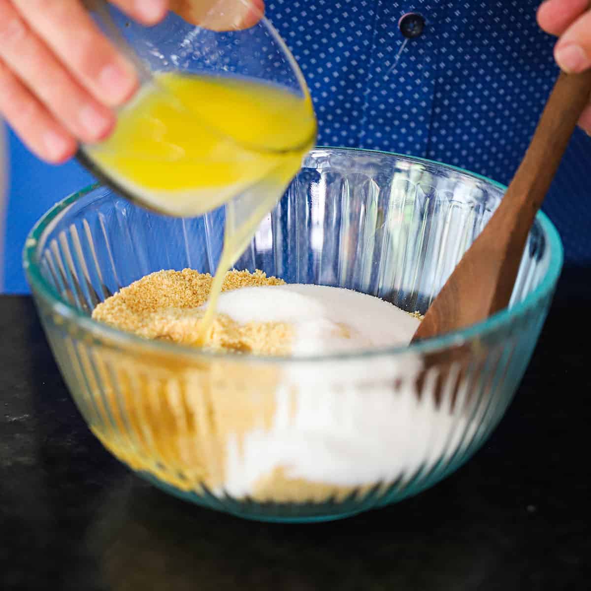 A person pouring melted butter in a glass bowl filled with crushed graham crackers, crushed vanilla wafers, and sugar.